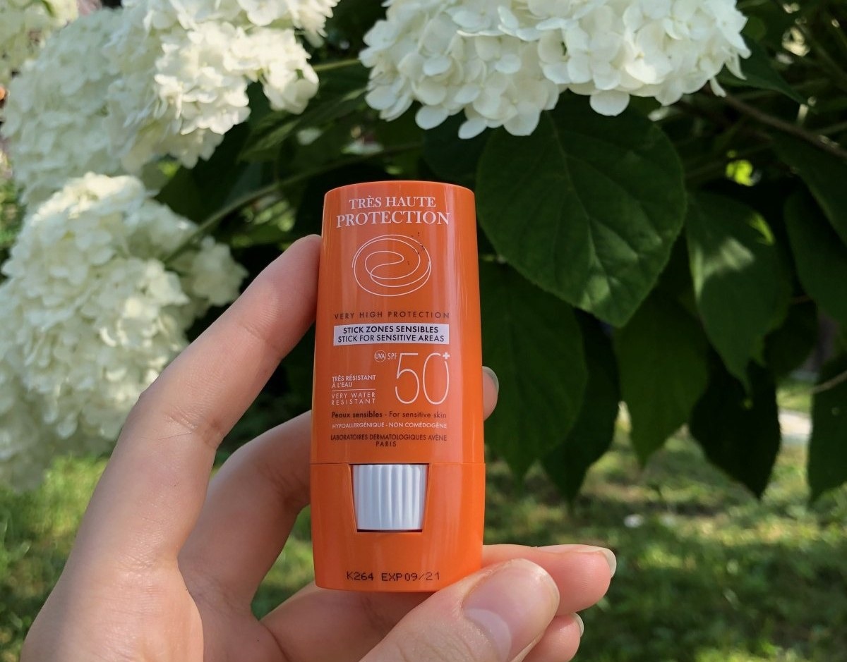 Avène Стик SPF 50 для лица — оранжевый стик для чувствительных зон в руке на фоне белых цветов