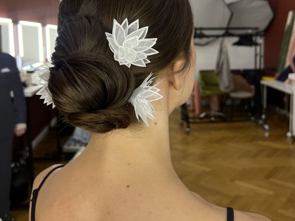 Низкий объёмный пучок на тёмных волосах, украшенный белыми цветами из органзы, вид сзади; снято в студии — свадебный образ