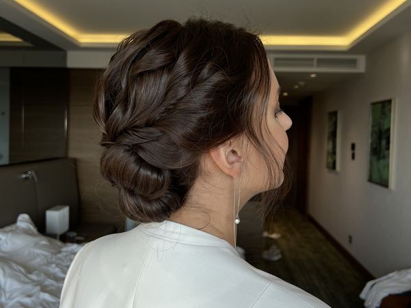 Вид сбоку на причёску: тёмные волосы собраны в низкий объёмный пучок с переплетёнными прядями, у лица выпущены тонкие пряди — макияж с выездом